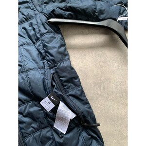 K&ouml;nnte beinhalten: Marineblaue Steppjacke mit Rei&szlig;verschluss und kleiner Tasche. Die Jacke hat ein Nike-Logo auf der Tasche und h&auml;ngt an einem schwarzen Kleiderb&uuml;gel. Die Jacke besteht aus einem gl&auml;nzenden, gesteppten Material.