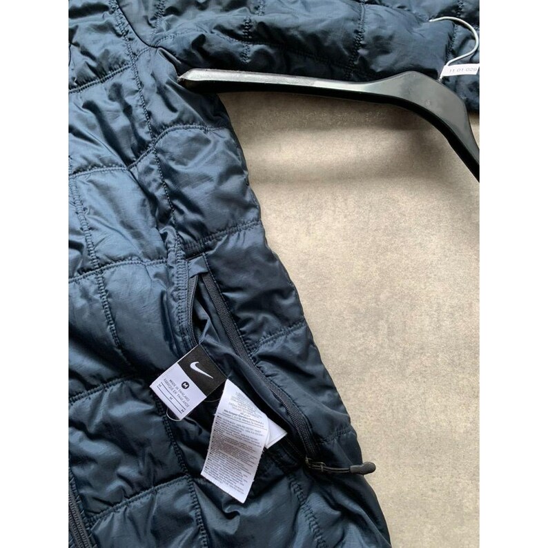 K&ouml;nnte beinhalten: Marineblaue Steppjacke mit Rei&szlig;verschluss und kleiner Tasche. Die Jacke hat ein Nike-Logo auf der Tasche und h&auml;ngt an einem schwarzen Kleiderb&uuml;gel. Die Jacke besteht aus einem gl&auml;nzenden, gesteppten Material.
