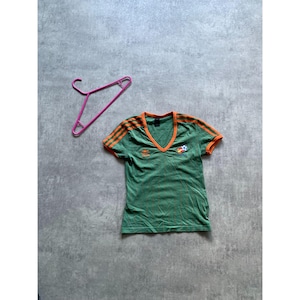 May include: Green v-neck t-shirt with orange stripes on the sleeves and collar. A soccer ball and emblem are on the chest. A pink hanger is next to the shirt.