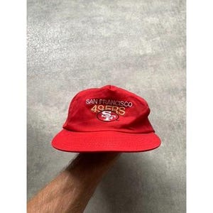 Peut inclure: Un chapeau de pêcheur rouge avec le texte "SAN FRANCISCO 49ERS" brodé en blanc et or. Le chapeau présente le logo des 49ers au centre. Le chapeau est fait d'une matière souple.