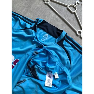 May include: Light blue athletic jersey with black accents and three black stripes on the sleeves. The jersey has a black collar and the Adidas logo is visible. A white hanger is in the background.