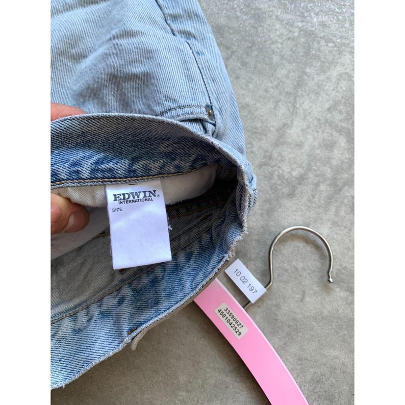 May include: Light blue denim garment with a white tag that reads "EDWIN INTERNATIONAL SIZE". A pink tag with the numbers "10 02 197" and other numbers is attached to a silver hanger.
