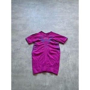 Puede incluir: Una camiseta magenta de manga corta con cuello redondo. La camiseta tiene un diseño texturizado en la parte delantera y alrededor de las mangas. El cuello presenta un patrón decorativo en azul, morado y naranja.