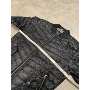 May include: Black quilted bomber jacket with a zipper closure, two front pockets, and a ribbed collar. A measuring tape is laid across the jacket. The jacket appears to be made of a lightweight material.