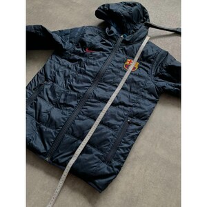 K&ouml;nnte beinhalten: Marineblaue Steppjacke mit Kapuze und durchgehendem Rei&szlig;verschluss. Mit rotem Nike-Logo und Barcelona-Teamwappen. Ein Ma&szlig;band liegt &uuml;ber der Jacke. Die Jacke hat zwei Rei&szlig;verschlusstaschen.