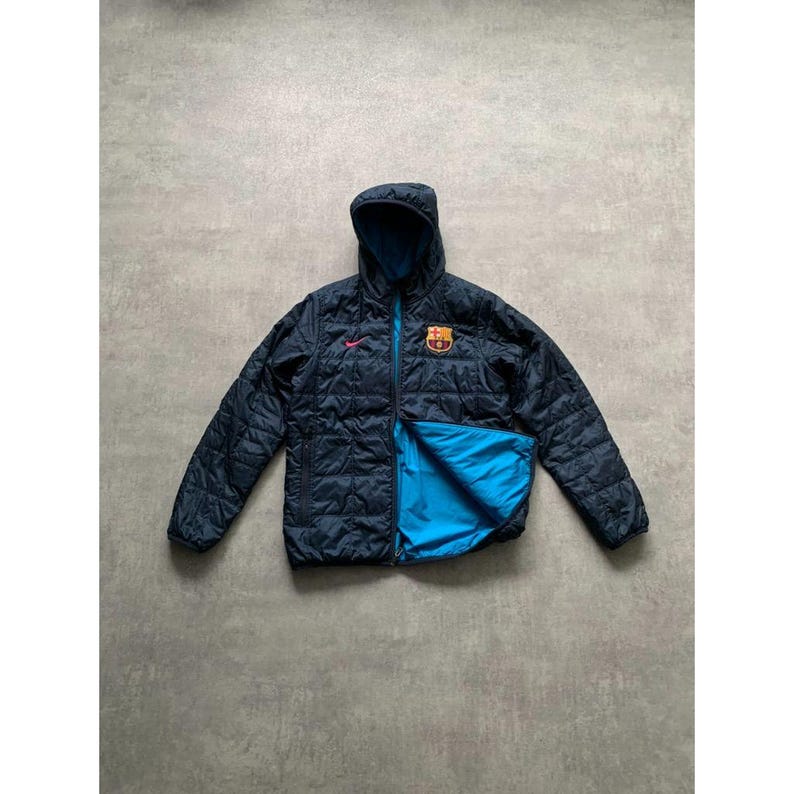 K&ouml;nnte beinhalten: Marineblaue Steppjacke mit Kapuze und hellblauem Innenfutter. Die Jacke hat einen Rei&szlig;verschluss, ein kleines rosa Logo und ein Team-Emblem auf der Brust. Die Jacke wird auf einer grauen Oberfl&auml;che pr&auml;sentiert.