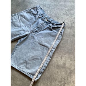 May include: Light blue denim shorts with a classic five-pocket design. A measuring tape is placed along the shorts, indicating the length. The shorts are made of a light-wash denim material.