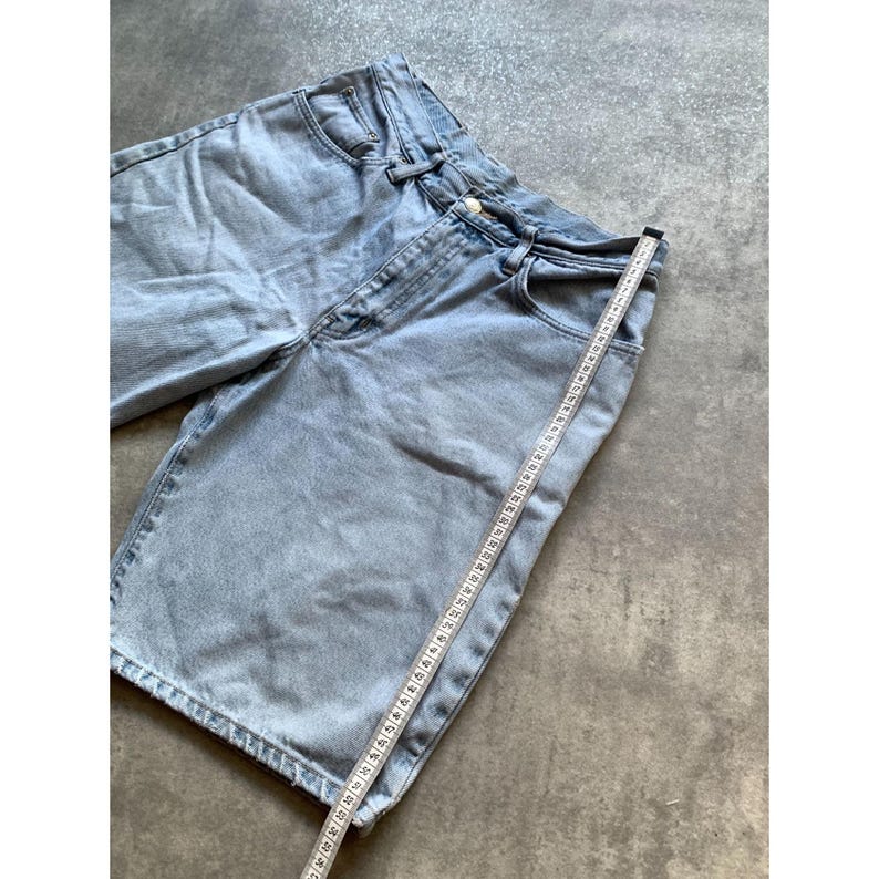 May include: Light blue denim shorts with a classic five-pocket design. A measuring tape is placed along the shorts, indicating the length. The shorts are made of a light-wash denim material.