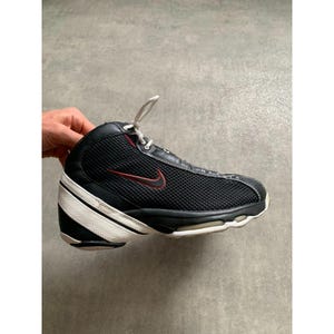 Puede incluir: Zapatilla deportiva negra y blanca con el logotipo rojo de la marca. La zapatilla tiene una parte superior de malla, cordones blancos y una suela blanca y negra. Es un modelo de ca&ntilde;a alta.