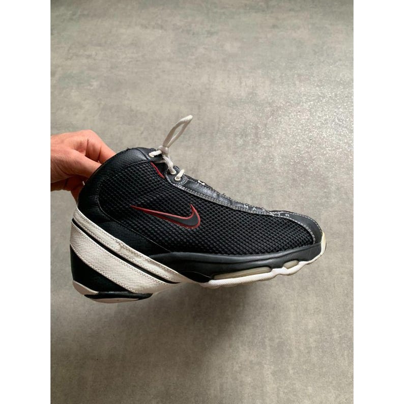 Puede incluir: Zapatilla deportiva negra y blanca con el logotipo rojo de la marca. La zapatilla tiene una parte superior de malla, cordones blancos y una suela blanca y negra. Es un modelo de ca&ntilde;a alta.