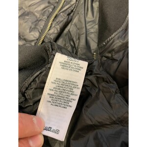 May include: Close-up of a black puffer jacket with a gold zipper. A white tag with text in multiple languages is visible. The tag lists the materials used in the jacket, including polyester, down, and feather.
