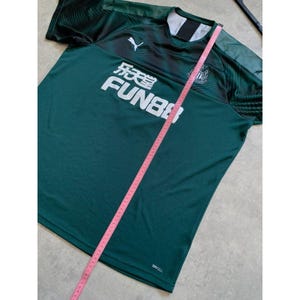 May include: A dark green athletic jersey with white lettering that reads "乐天 FUN88". The jersey has a white Puma logo and a crest on the upper left chest. A pink measuring tape is laid across the jersey.