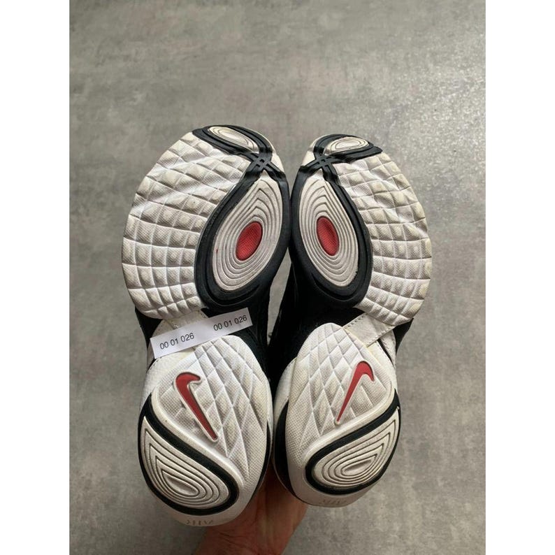 Puede incluir: Par de zapatillas deportivas negras y blancas con detalles rojos. Las suelas son blancas con un patr&oacute;n texturizado y un logotipo Nike rojo. Las zapatillas tienen un dise&ntilde;o en blanco y negro con un detalle circular rojo.