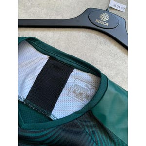 May include: Close-up of a dark green Puma athletic jersey. The jersey has a black collar and a white mesh panel. The Puma logo is visible on a tag. The jersey is hanging on a black hanger with the Bosca logo.