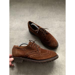 Puede incluir: Un par de zapatos brogue de ante marrón con un diseño clásico. Los zapatos presentan perforaciones decorativas y cordones marrones. Las suelas son de color marrón oscuro. Hay hormas en cada zapato.