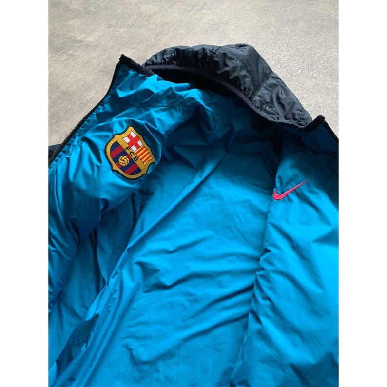 K&ouml;nnte beinhalten: Wendejacke in Marineblau und T&uuml;rkis mit dem FC Barcelona-Teamlogo und einem pinkfarbenen Nike-Swoosh. Die Jacke hat eine Kapuze und ein gestepptes Design.