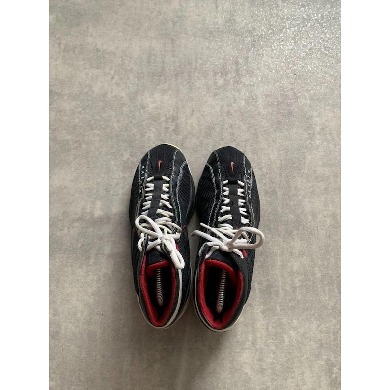 Puede incluir: Par de zapatillas deportivas negras con cordones blancos y detalles rojos. Las zapatillas tienen una parte superior de malla y un peque&ntilde;o logotipo rojo de Nike. Las suelas son blancas y el interior es rojo.