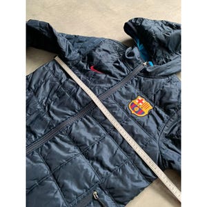 K&ouml;nnte beinhalten: Marineblaue, gesteppte Jacke mit Kapuze, rotem Nike-Logo und Barcelona-Teamwappen. Die Jacke hat einen Rei&szlig;verschluss und ein Rautenmuster. Ein Ma&szlig;band liegt &uuml;ber der Jacke.