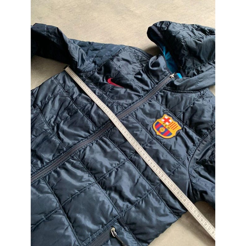 K&ouml;nnte beinhalten: Marineblaue, gesteppte Jacke mit Kapuze, rotem Nike-Logo und Barcelona-Teamwappen. Die Jacke hat einen Rei&szlig;verschluss und ein Rautenmuster. Ein Ma&szlig;band liegt &uuml;ber der Jacke.