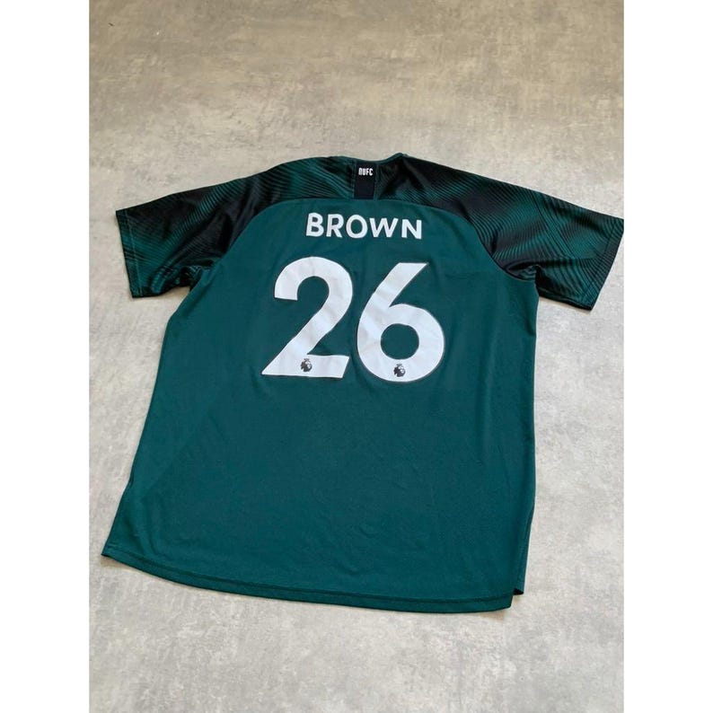 May include: A dark green athletic jersey with the name "BROWN" and the number "26" in white. The sleeves are black with a green geometric pattern. The jersey is made of a breathable material.