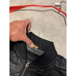 May include: Black quilted bomber jacket with a black ribbed collar and a silver zipper. The jacket has the "True Religion" logo on a tag inside the collar. The jacket is hanging on a red hanger.