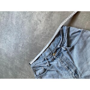 May include: Light blue denim jeans with a button closure and pockets. A white measuring tape is laid across the waistband. The jeans are on a gray surface.