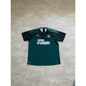 May include: A dark green short-sleeved soccer jersey with the text "乐天" and "FUN88" in white. The jersey has a black and green design on the shoulders and sleeves, and a team crest on the chest.