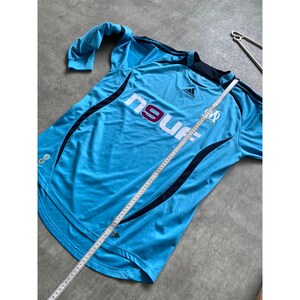 May include: Light blue Adidas soccer jersey with the text "ngur" and a team logo. The jersey has black accents and a crew neck. A measuring tape is laid across the jersey.