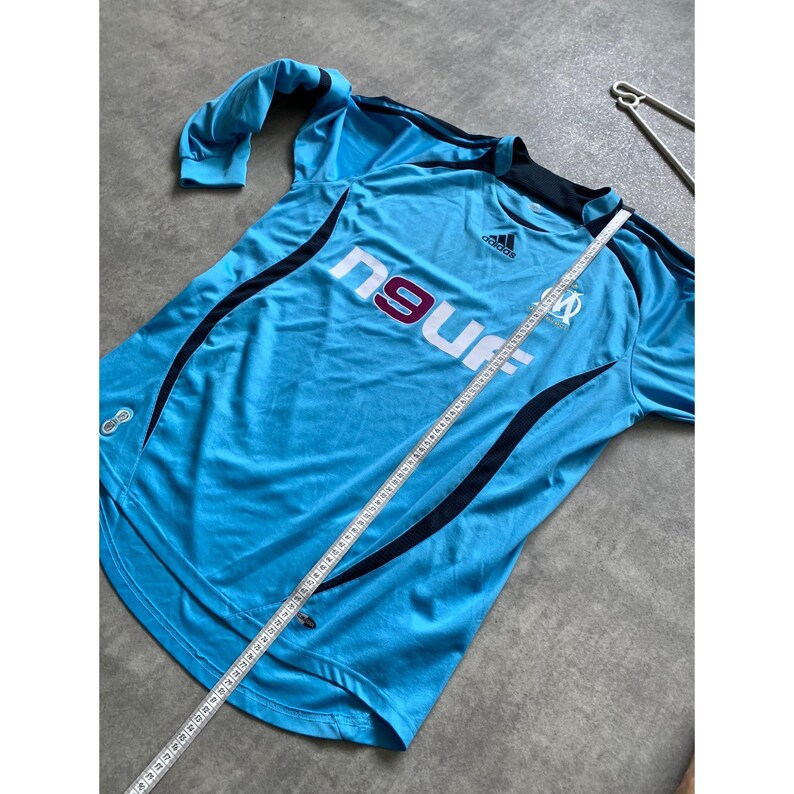 May include: Light blue Adidas soccer jersey with the text "ngur" and a team logo. The jersey has black accents and a crew neck. A measuring tape is laid across the jersey.