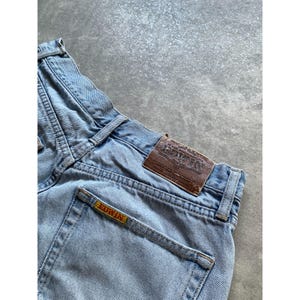 May include: Light blue denim jeans with a faded wash. The jeans feature a brown leather patch with the brand name "EDWIN" and a yellow label with the brand name on the back pocket. The jeans have a classic five-pocket design.