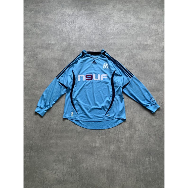 May include: Light blue long-sleeved soccer jersey with black accents. The jersey features the word "neuf" in white, a logo on the chest, and black stripes on the shoulders. The jersey is made of a lightweight material.