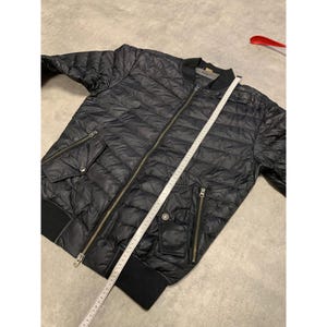 May include: Black quilted bomber jacket with a zipper closure, two front pockets, and a ribbed collar and cuffs. A measuring tape is laid across the jacket. The jacket is approximately 28 inches long.