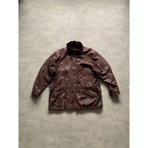 Puede incluir: Chaqueta de cuero marrón con cuello alto, cierres de botón y cintura con cordón. La chaqueta tiene múltiples bolsillos y un forro acolchado. El cuero parece desgastado, dándole un aspecto vintage. La chaqueta mide aproximadamente 76 cm de largo.