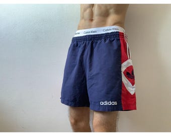 May include: Navy blue Adidas athletic shorts with a red and white panel on the right side. The panel features the Adidas logo and three white stripes. The word "adidas" is printed in white on the left leg.