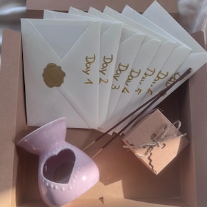 Könnte beinhalten: Geschenkset in einer braunen Box, mit cremefarbenen Umschlägen mit der Aufschrift „Tag 1“ bis „Tag 6“ in Gold. Enthält einen rosa Keramik-Öldiffusor mit herzförmigem Ausschnitt, Räucherstäbchen und eine kleine braune Geschenkbox.