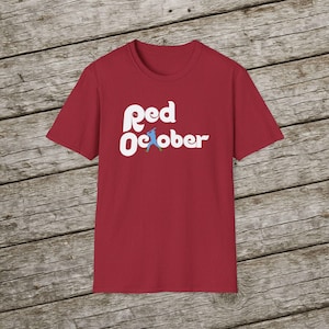 May include: A red t-shirt featuring the white text "Red October" in a decorative font. A small blue and green graphic is positioned between the words. The shirt is shown on a wooden background.