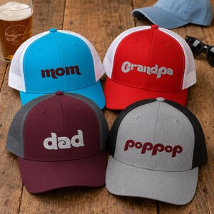 May include: Four trucker hats with mesh backs. One is bright blue with 'mom' in maroon, one is red with 'Grandpa' in white, one is maroon with 'dad' in white, and one is heather gray with 'poppop' in maroon. Each hat features a structured front panel.