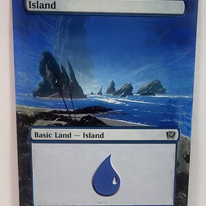 Puede incluir: Una carta de Islandia azul y blanca de un juego de cartas coleccionables. La carta presenta un paisaje marino con islas rocosas, un símbolo de gota de agua azul y el texto "Island" y "Basic Land - Island".