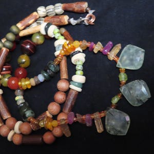 Op de afbeelding: Een handgemaakte ketting met een verscheidenheid aan kralen in bruin, groen, rood en geel. De ketting bevat houten kralen, edelstenen en facetgeslepen groene stenen, wat een uniek en aards ontwerp creëert. De ketting wordt gepresenteerd tegen een zwarte achtergrond.