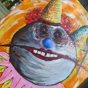 May include: A painting of a clown's face with a blue nose, red lips, and a brown and gold hat. The clown's face is painted in shades of grey and white, with red accents around the eyes and mouth. The background is orange.