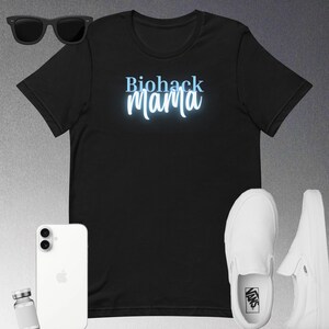 May include: Black t-shirt with the text "Biohack Mama" in light blue, stylized script. The image also includes black sunglasses, a white smartphone, a small vial, and white slip-on sneakers.