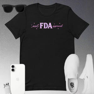 Peut inclure: T-shirt noir avec le texte "not FDA approved" en rose. L'image comprend également des lunettes de soleil noires, un smartphone blanc, un petit flacon et des baskets Vans blanches.