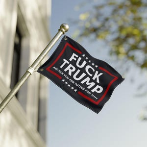 May include: A black flag with the words "FUCK TRUMP" in white, outlined in red. The flag also includes the text "AND FUCK YOU FOR VOTING FOR HIM" in white. The flag is attached to a gold pole.