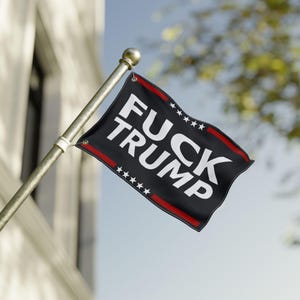 May include: Black flag with white text reading "FUCK TRUMP". The flag has red trim and white stars. It is mounted on a gold pole and is waving in the wind against a blue sky.