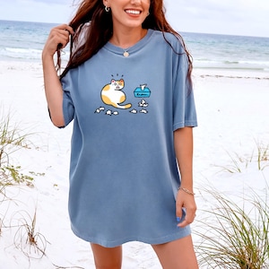 May include: A blue t-shirt with a graphic of a surprised cat next to a tissue box and scattered tissues. The cat is white and orange. The shirt is a solid light blue color.