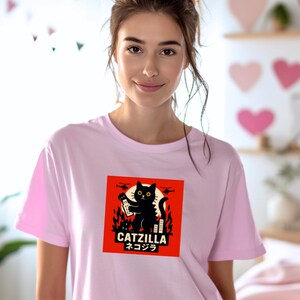 May include: A light pink t-shirt featuring a graphic of a black cat with yellow eyes, holding a bottle, set against a red background. The design includes the text "CATZILLA" and Japanese characters. The shirt is being worn.