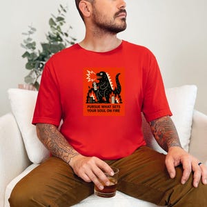 May include: Red t-shirt featuring a black illustration of a Godzilla-like creature breathing fire, with the text "PURSUE WHAT SETS YOUR SOUL ON FIRE" in a rectangular orange box. The shirt is short-sleeved and worn by a person.