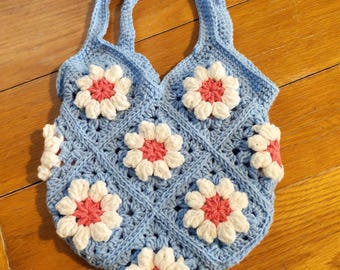 Puede incluir: Un bolso tote de ganchillo azul claro con aplicaciones de flores blancas y rosas. El bolso presenta un patrón de diamantes y dos asas. El bolso mide aproximadamente 25 cm de ancho.