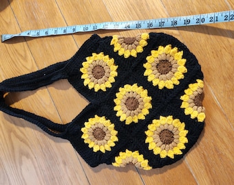 Puede incluir: Bolso tote de crochet negro con un estampado de girasoles. El bolso tiene un asa larga y mide aproximadamente 56 cm de ancho. Los girasoles son amarillos y marrones, sobre un fondo negro.