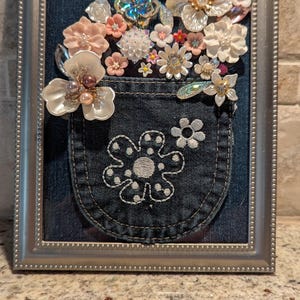 May include: A framed artwork featuring a dark blue denim pocket mounted on a dark blue denim background. The pocket is adorned with white embroidered flower outlines and a collection of various artificial flowers in white, pink, and iridescent tones.